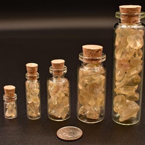 May include: Five clear glass bottles with cork stoppers, each filled with light yellow-brown gemstones. The bottles vary in size, with the tallest one containing the most stones. A quarter is in front of the bottles.
