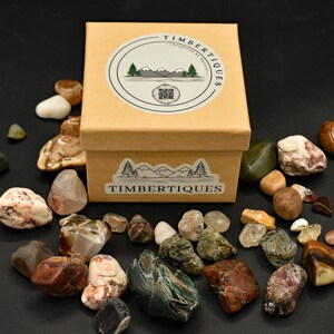 May include: A collection of polished stones in various colors and textures surrounds a small, tan box. The box is labeled "TIMBERTIQUES" with a mountain and tree logo. The stones include shades of brown, green, white, and red.