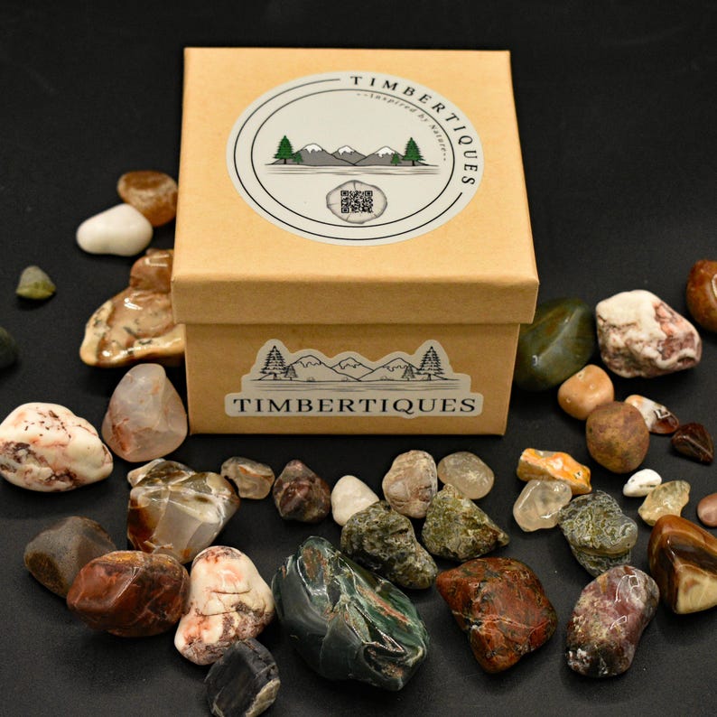 May include: A collection of polished stones in various colors and textures surrounds a small, tan box. The box is labeled "TIMBERTIQUES" with a mountain and tree logo. The stones include shades of brown, green, white, and red.