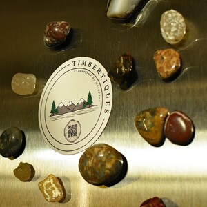May include: A collection of polished stones in various colors, including brown, tan, and gray, arranged around a white oval magnet. The magnet features a mountain and tree design with the text "TIMBERTIQUES - Inspired by Nature -" and a QR code.