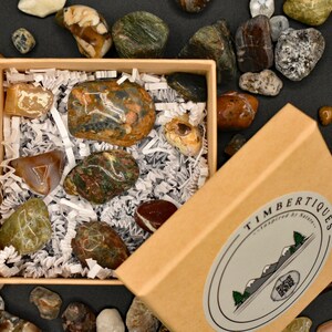 May include: A collection of polished stones in various colors and patterns, presented in a kraft paper box with shredded paper. The box has a label with the text "TIMBERTIQUES - Inspired by Nature". Additional stones are scattered around the box.