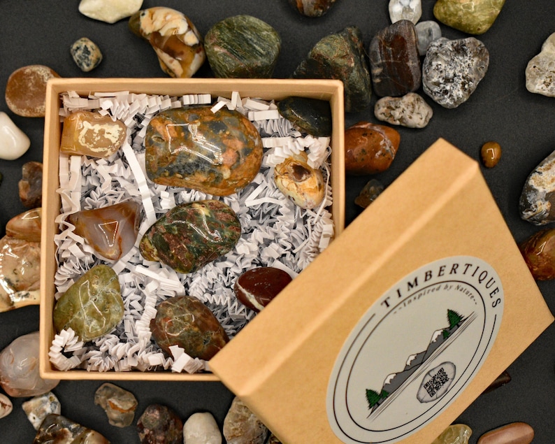 May include: A collection of polished stones in various colors and patterns, presented in a kraft paper box with shredded paper. The box has a label with the text "TIMBERTIQUES - Inspired by Nature". Additional stones are scattered around the box.