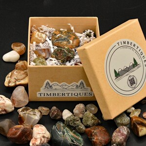 May include: A collection of polished stones in various colors and patterns, displayed in and around a kraft paper box. The box is labeled "TIMBERTIQUES" and features a mountain and tree logo. The stones vary in size and shape, with some inside the box and others scattered around it.