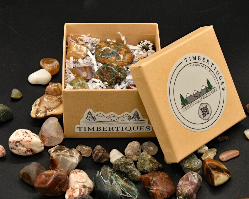 May include: A collection of polished stones in various colors and patterns, displayed in and around a kraft paper box. The box is labeled "TIMBERTIQUES" and features a mountain and tree logo. The stones vary in size and shape, with some inside the box and others scattered around it.