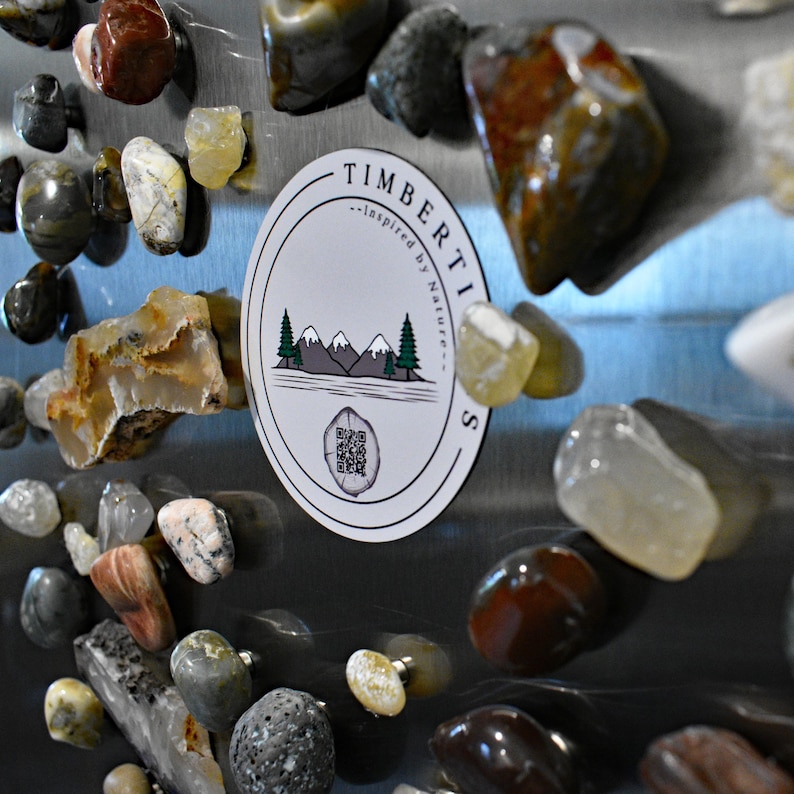 May include: A collection of polished stones in various colors, including brown, gray, and yellow, are attached to a metallic surface. A white oval sticker with the text "TIMBERTICS" and a nature-inspired design is also affixed.