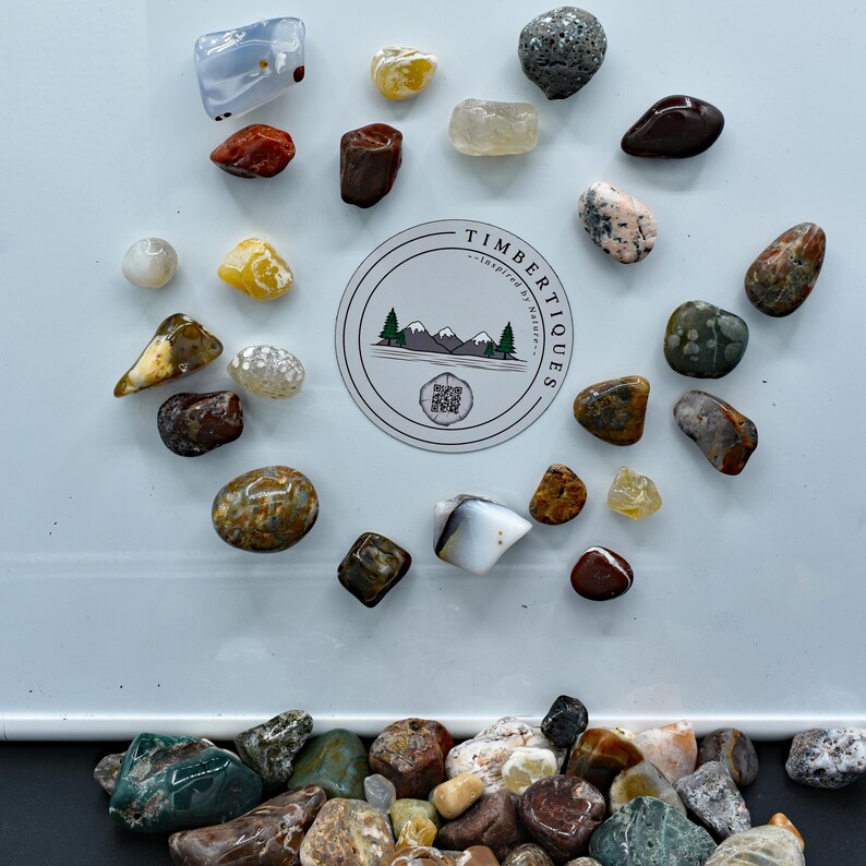 May include: A collection of polished gemstones in various shapes and colors, including red, brown, yellow, and gray. A circular sticker with the text "TIMBERTIQUES - inspired by nature" is in the center. The gemstones are scattered on a white surface.