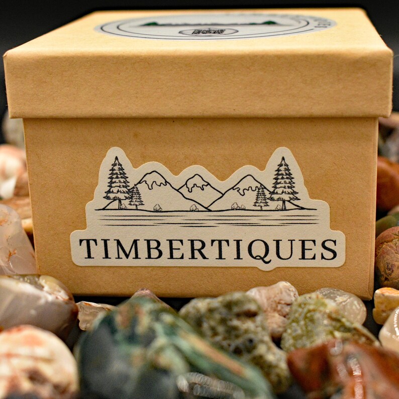May include: A tan cardboard box with a sticker featuring a mountain and tree design, with the word "TIMBERTIQUES" below. The box is surrounded by colorful rocks.