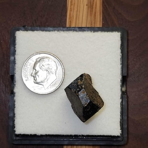 May include: A black, faceted, crystal-like stone, approximately the size of a US dime, displayed on a white background.