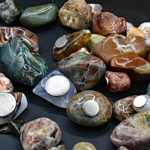 May include: A collection of polished, colorful stones with embedded silver magnets. The stones vary in size and shape, displaying a range of colors including green, brown, red, and gray. The magnets are circular and affixed to the flat surfaces of the stones.