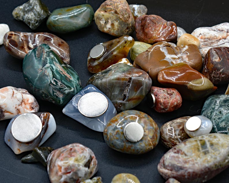 May include: A collection of polished, colorful stones with embedded silver magnets. The stones vary in size and shape, displaying a range of colors including green, brown, red, and gray. The magnets are circular and affixed to the flat surfaces of the stones.