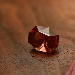 May include: A faceted, reddish-brown gemstone with a geometric shape. The gem has a reflective surface and sits on a dark wood surface. The gem's facets catch the light, creating highlights and shadows.