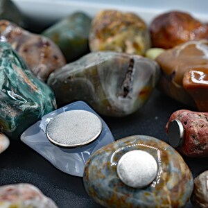 May include: A collection of polished stones in various colors, including green, brown, and red. Each stone has a flat, circular magnet attached to it. The stones vary in size and shape, with a natural, organic appearance.