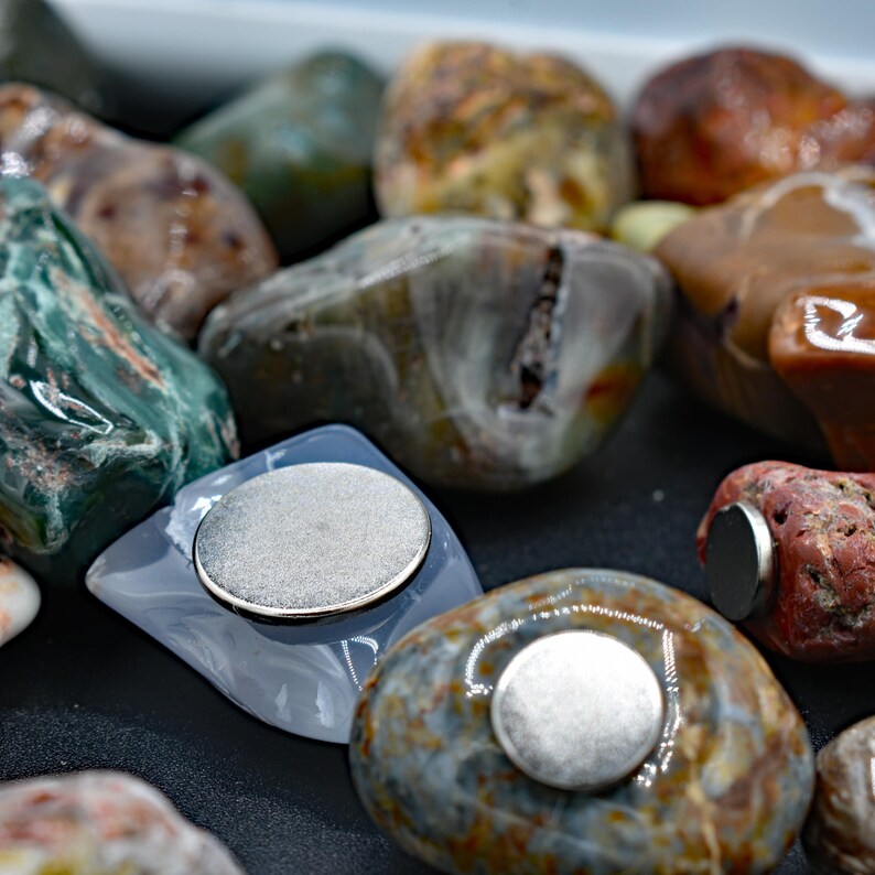 May include: A collection of polished stones in various colors, including green, brown, and red. Each stone has a flat, circular magnet attached to it. The stones vary in size and shape, with a natural, organic appearance.
