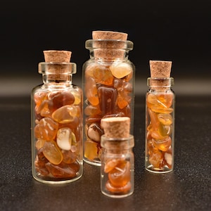 May include: Four clear glass bottles with cork stoppers, each filled with polished, amber-colored gemstones. The bottles vary in size, with the gemstones visible through the glass. The background is a dark, solid color.