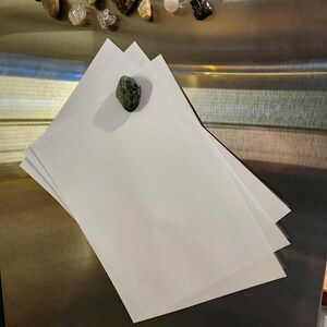 May include: A stack of white paper sheets with a green and brown stone magnet. Assorted gemstones are visible in the background. The paper is on a metallic surface.