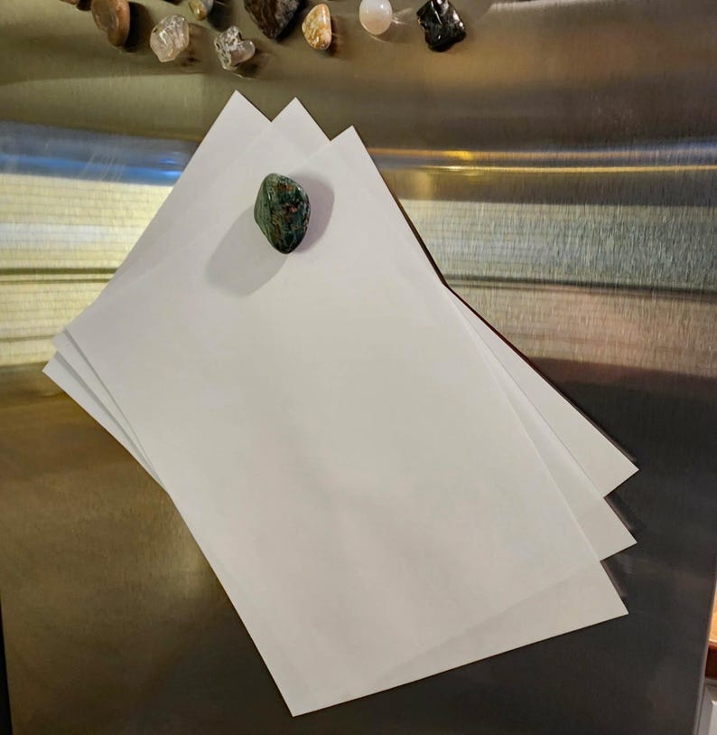 May include: A stack of white paper sheets with a green and brown stone magnet. Assorted gemstones are visible in the background. The paper is on a metallic surface.