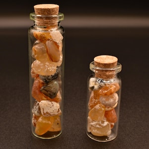 May include: Two small glass bottles filled with polished gemstones. The larger bottle contains a variety of stones in shades of orange, tan, and gray. The smaller bottle also contains similar stones. Both bottles have cork stoppers.