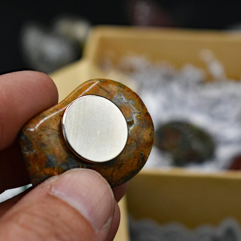 May include: A natural stone magnet with a polished, rounded shape. The stone features a mix of brown, orange, and gray hues. A silver-colored, circular magnet is affixed to the stone's surface.