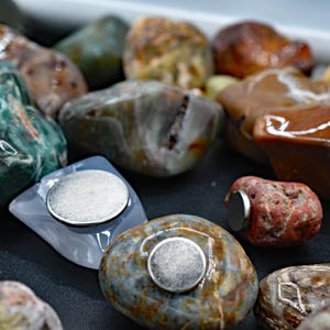 May include: A collection of polished, colorful stones, some with attached silver magnets. The stones vary in color, including shades of green, brown, and gray. The magnets are round and metallic, affixed to the flat surfaces of the stones.