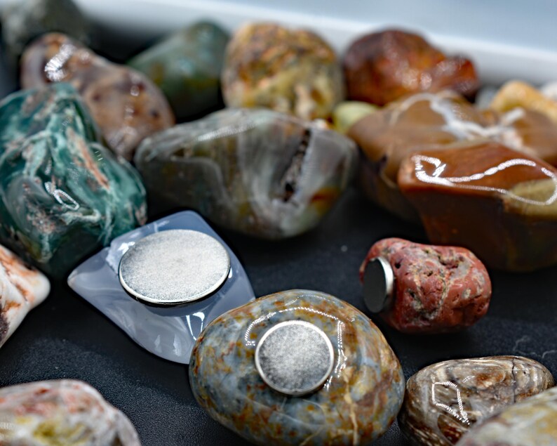 May include: A collection of polished, colorful stones, some with attached silver magnets. The stones vary in color, including shades of green, brown, and gray. The magnets are round and metallic, affixed to the flat surfaces of the stones.