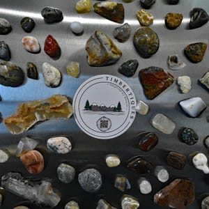 May include: A collection of polished stones in various shapes, sizes, and colors, including red, white, yellow, and gray. A circular sticker with the text "TIMBERTIQUES - inspired by Nature" and a mountain and tree graphic is also present.