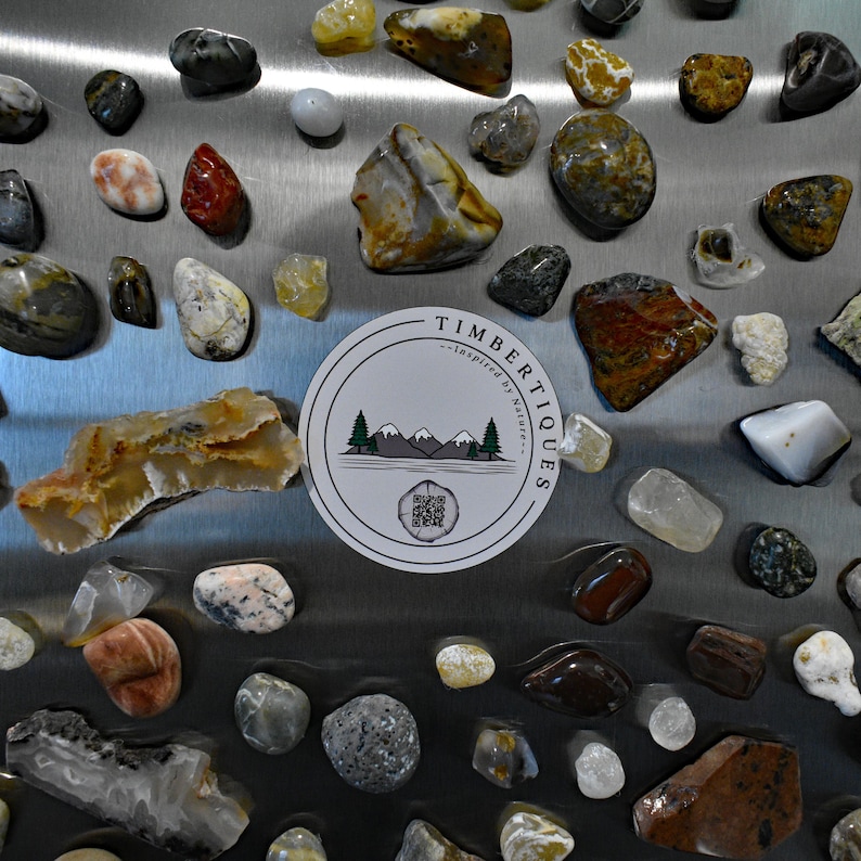 May include: A collection of polished stones in various shapes, sizes, and colors, including red, white, yellow, and gray. A circular sticker with the text "TIMBERTIQUES - inspired by Nature" and a mountain and tree graphic is also present.