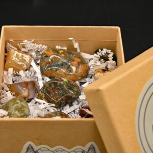 May include: A partially open, tan cardboard box filled with various polished stones. The stones are in shades of brown, green, orange, and white, nestled in white paper filler. The box has a mountain and tree sticker on the side.