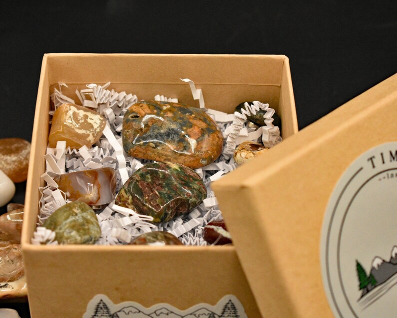 May include: A partially open, tan cardboard box filled with various polished stones. The stones are in shades of brown, green, orange, and white, nestled in white paper filler. The box has a mountain and tree sticker on the side.
