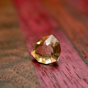May include: A faceted, golden-yellow gemstone with multiple reflective surfaces. The gem is set against a wooden surface with alternating bands of brown and red-purple hues, creating a contrasting backdrop. The gem's shape is irregular, with a flat top and angled sides.