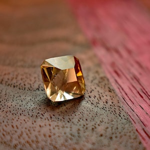 May include: A faceted, golden-yellow gemstone with a square cushion cut. The gem is set against a textured, brown surface with a blurred pink and red background, creating a contrast of colors and textures. The gem's facets reflect light.