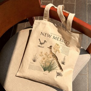 Könnte beinhalten: Eine beigefarbene Tragetasche aus Segeltuch mit einem botanischen und tierischen Aufdruck. Das Design enthält den Text New Mexico, Bird & Bloom Collection, Greater Roadrunner und Yucca sowie Illustrationen von Roadrunnern und Yuccablüten.