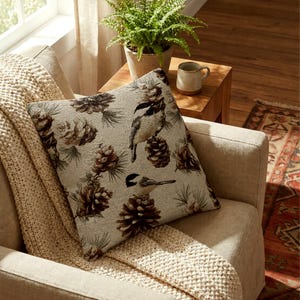 May include: A square throw pillow featuring a woven pattern of brown pinecones, green pine needles, and black and white chickadee birds on a light beige background. The fabric has a textured appearance with a repeating nature-inspired motif.