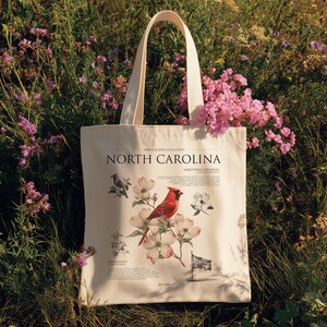 May include: Cream-colored tote bag featuring a red cardinal bird and dogwood flowers, with the text "NORTH CAROLINA" and "NORTHERN CARDINAL". The bag has a vintage botanical illustration style and a cream-colored handle.