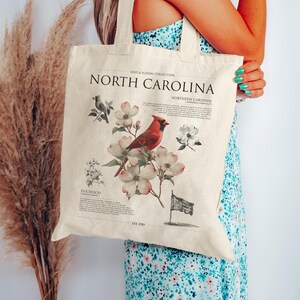 May include: Cream-colored tote bag featuring a North Carolina design from the Bird & Bloom Collection. The design includes a red cardinal, dogwood flowers, and the text "North Carolina." The bag has a vintage aesthetic and sturdy handles.
