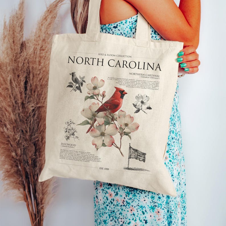 May include: Cream-colored tote bag featuring a North Carolina design from the Bird & Bloom Collection. The design includes a red cardinal, dogwood flowers, and the text "North Carolina." The bag has a vintage aesthetic and sturdy handles.