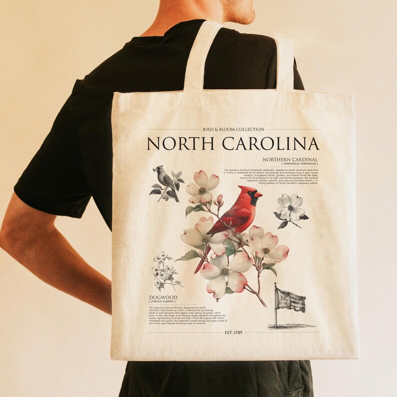 May include: Cream-colored tote bag featuring a North Carolina design. The design includes a red cardinal, dogwood flowers, and the text "North Carolina." The bag has two white straps.