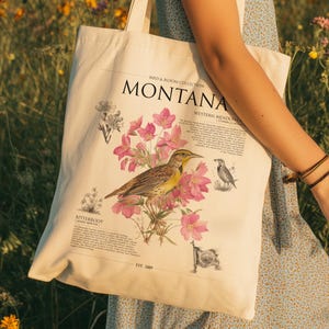 Puede incluir: Bolso tote de color crema con una ilustración de pájaro y flores. El diseño incluye un pájaro con plumas amarillas y marrones, flores rosas y la palabra "MONTANA". La bolsa también tiene el texto "Bird & Bloom Collection" y "Western Meadow".