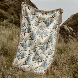 May include: A woven throw blanket featuring a repeating pattern of light blue flowers, green leaves, and small birds on a cream-colored background. The rectangular blanket has a textured fringe border along all four edges of the fabric.
