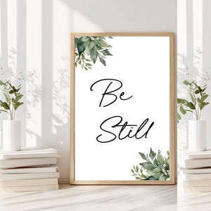 Può includere: Una stampa incorniciata bianca con un disegno di foglie verdi in alto e in basso. La stampa dice "Be Still" in scrittura corsiva nera.