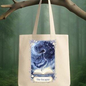 Può includere: Borsa tote beige con un'illustrazione ad acquerello di una donna in abito blu in piedi su un libro aperto, sullo sfondo di un cielo stellato. Il testo "The Escapist" è sotto l'immagine, incorniciato da elementi floreali.
