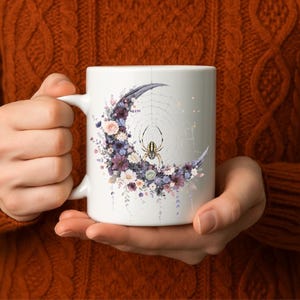 May include: White ceramic mug featuring a crescent moon design with a spider web and a spider. The moon is adorned with purple, pink, and white flowers. The mug is held by a person wearing an orange sweater.