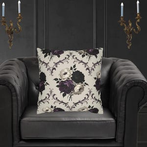 May include: Square cushion with a floral pattern featuring dark purple, white, and black roses on a cream background. The cushion has a damask-style design with swirling vines. The cushion is placed on a black leather armchair.
