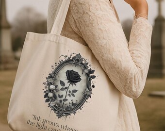 May include: A cream-colored tote bag featuring a black rose illustration within an ornate frame. The design includes floral accents and the quote, "Ink grows where the light can't reach." The bag is held by a person wearing a cream-colored dress.