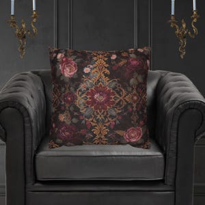 Op de afbeelding: Een vierkant sierkussen met een bloemenpatroon in bordeaux, goud en roze tinten op een donkere achtergrond. Het kussen ligt op een zwarte leren fauteuil, wat een gotische of vintage uitstraling creëert.