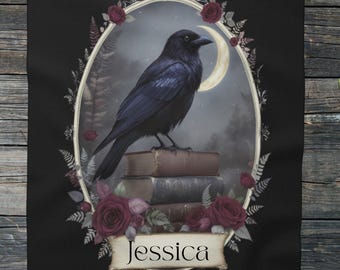May include: Black throw blanket featuring a gothic-style illustration. A black raven perches atop stacked books within an oval frame, surrounded by roses and ferns. The name "Jessica" is displayed on a banner below the frame.
