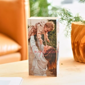 Op de afbeelding: Een houten fotokubus met zes zijden, elke zijde toont een andere foto van een moeder en kind. De foto's zijn gedrukt in warme tinten en tonen de moeder die het kind in de lucht houdt. De kubus is versierd met de tekst "lovegiftedom studio" in roze.
