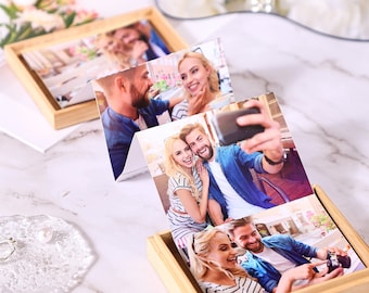Puede incluir: Un conjunto de tres fotografías impresas con una pareja sonriente, expuestas en una pequeña caja de madera de color claro. Las fotos muestran a la pareja en varias poses, incluyendo un selfie. La caja está sobre una superficie de mármol.