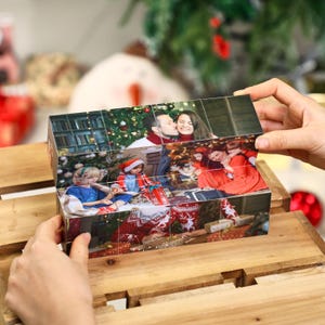 May include: A personalized photo cube featuring family photos. The cube is made of multiple blocks, each displaying a different image. The photos show people in a festive setting, with a Christmas tree in the background. The cube is being held on a wooden surface.