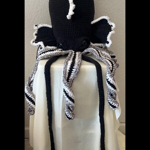 May include: A handmade crochet octopus hat in black with white accents. The hat features a large head with white trim and long, striped tentacles in black, white, and gray. The octopus is displayed on a white surface.