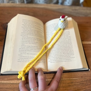 May include: A handmade crochet chicken bookmark with a white body, red comb, and yellow legs. The bookmark is placed in an open book, adding a whimsical touch to reading. The book is open to page 151.